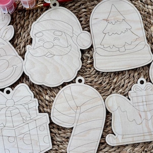 May include: A collection of unfinished wooden Christmas ornaments in various shapes, including Santa Claus, a reindeer, a snowman, a candy cane, a gift box, a snow globe with a tree, and a sleigh. Each ornament has a pre-drilled hole for hanging.