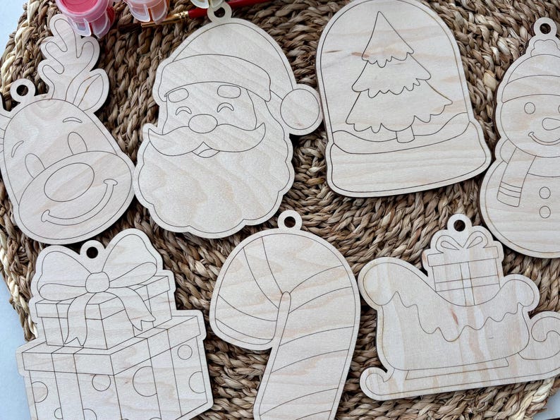 May include: A collection of unfinished wooden Christmas ornaments in various shapes, including Santa Claus, a reindeer, a snowman, a candy cane, a gift box, a snow globe with a tree, and a sleigh. Each ornament has a pre-drilled hole for hanging.