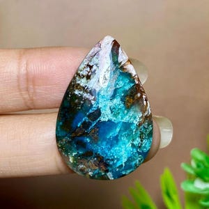 Puede incluir: Una gema en forma de lágrima con una vibrante mezcla de colores. La piedra presenta una llamativa combinación de azules profundos, turquesas y marrones, con acentos blancos y marrón claro. La superficie pulida de la piedra refleja la luz.