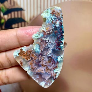 May include: A polished agate slice featuring a translucent, pale lavender base with intricate, branching inclusions in shades of deep rust-red, copper, and vibrant turquoise. The stone has an irregular, organic shape with a smooth, glossy surface.