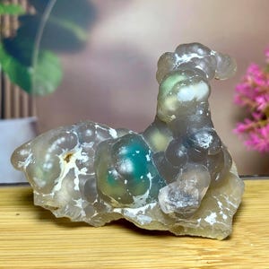 Puede incluir: Un espécimen mineral natural único con una forma orgánica y burbujeante. La piedra presenta formaciones redondeadas translúcidas en tonos de gris, verde y blanco, que descansan sobre una base de color amarillo claro. El espécimen se exhibe sobre una superficie de madera.