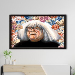May include: A framed portrait of a man with white hair and glasses, set against a floral background. The artwork is displayed on a white wall above a modern wooden cabinet, with a blue armchair and ottoman in the foreground.