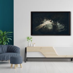 May include: A framed photograph of a woman in a white dress lying in a dark forest setting. The photograph is hanging on a white wall above a wooden and white media console.