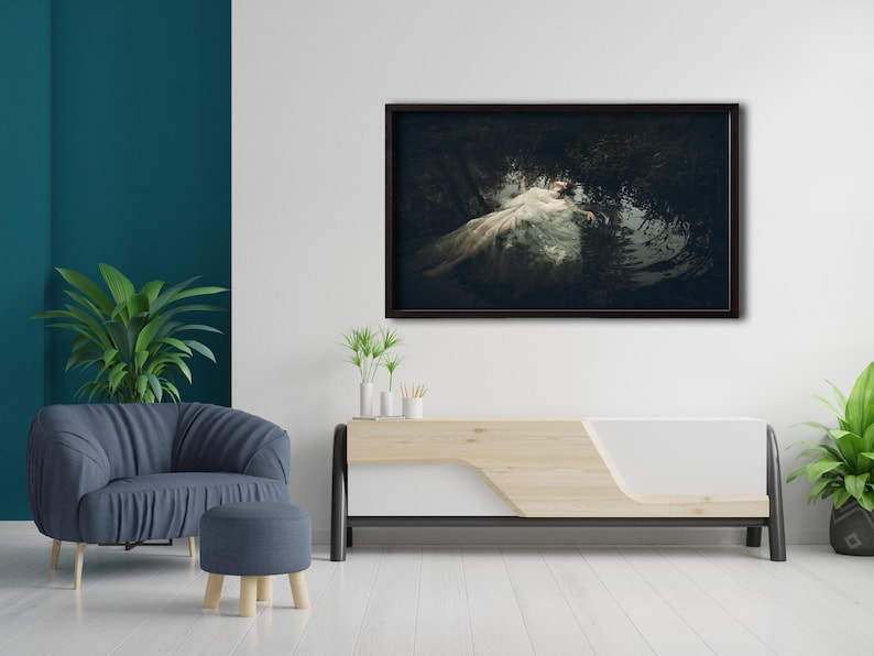 May include: A framed photograph of a woman in a white dress lying in a dark forest setting. The photograph is hanging on a white wall above a wooden and white media console.
