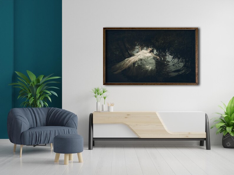 May include: A framed photograph of a woman in a white dress lying in a dark forest setting. The photograph is hanging on a white wall above a light wood and white entertainment center.