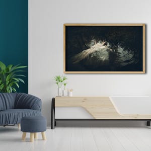 May include: A framed photograph of a woman in a white dress lying in a dark forest setting. The photograph is hanging on a white wall above a wooden and white media console.