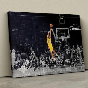 May include: A black and white photo of a basketball player in mid-air, about to dunk the ball. The player is wearing a yellow and purple jersey with the number 24 on it. The player is surrounded by other players and fans, all in black and white. The score on the scoreboard is 0-4.