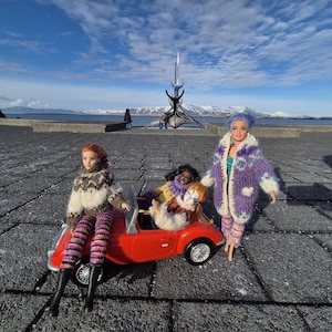 Könnte beinhalten: Ein rotes Cabrio mit vier Puppen, eine Puppe trägt einen braun-weißen Pullover, gestreifte Hosen und schwarze Stiefel. Eine andere Puppe trägt einen lila Mantel und eine Hose. Die Puppen befinden sich auf einem gepflasterten Bereich mit einer Skulptur und Bergen im Hintergrund.