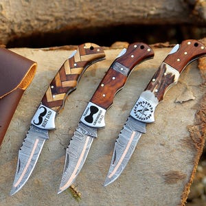 May include: Three pocket knives with different wood handles and engraved designs. The knives have a damascus steel blade and are displayed on a wooden surface. The knives are accompanied by a brown leather pouch with the initials "BCK" engraved on it.