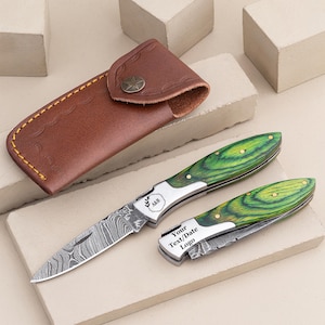 May include: A green and silver pocket knife with a wood handle and a leather sheath. The knife has a damascus blade and is engraved with the text "A&B" and "Your Text/Date Logo".