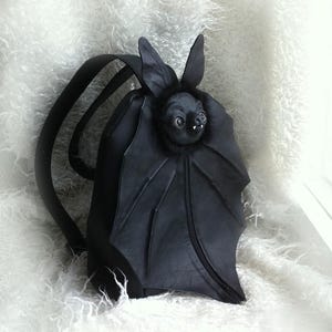 May include: A black backpack shaped like a bat, featuring a sculpted bat head with pointed ears, small white fangs, and large wings that extend across the front. The bag includes two shoulder straps and is constructed from a smooth, matte material.