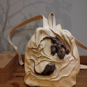 Könnte beinhalten: Cremefarbener Lederrucksack mit einem 3D-Skulptur-Design von zwei Eidechsen. Die Eidechsen sind braun und scheinen aus der strukturierten Oberfläche der Tasche herauszukommen. Der Rucksack hat verstellbare Träger und einen oberen Griff.