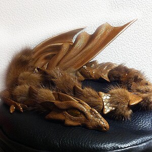 May include: A brown dragon figurine with textured leather wings and scales, and fluffy fur accents. The dragon is lying down on a black surface, with its head and tail curved. The wings are spread out.