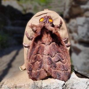 Puede incluir: Una mochila de cuero color beige con un diseño único. La mochila presenta un adorno de piel marrón y un adorno decorativo con forma de insecto con lentes naranjas. La mochila está colocada sobre un fondo natural al aire libre.