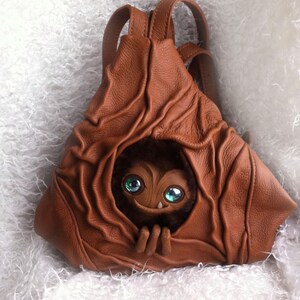 May include: A brown leather backpack with a furry creature with large green eyes peeking out of the top opening.