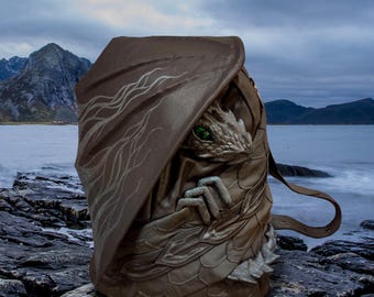 May include: A brown leather dragon backpack with green eyes and a detailed design. The backpack is sitting on a rocky surface near a body of water.