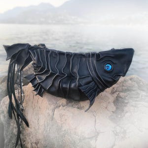 May include: A black leather handbag shaped like a fish, with a textured body and fins. The bag features a blue eye and a long, beaded tail. The bag is designed to resemble a fish.