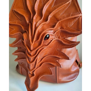 May include: A cognac-coloured leather backpack featuring a three-dimensional, layered, leaf-like or scale-like design on the front panel. The structure includes a prominent, pointed central element and a single visible dark, glossy eye-like accent.