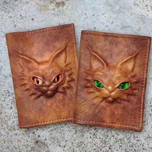 Puede incluir: Dos fundas rectangulares para pasaporte de cuero marrón con caras de gato en relieve. Un gato tiene ojos de color naranja brillante, mientras que el otro tiene ojos de color verde vibrante. El cuero tiene un acabado texturizado con costuras.