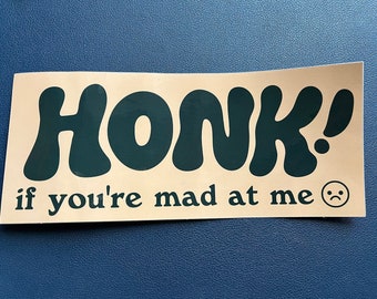 Può includere: Un adesivo beige con testo nero che dice "HONK! if you're mad at me" e un'emoji di faccina triste in bianco e nero.