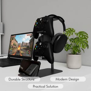 May include: Black gaming controller and headset stand. The stand holds two controllers, a headset, and a smartphone. The stand is black and has the words "Durable Structure", "Modern Design", and "Practical Solution" on the bottom.