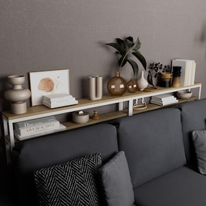 May include: A modern, two-tiered shelf unit with a white metal frame and light wood shelves. The shelves are filled with decorative items, including vases, books, and framed artwork. The unit is positioned above a gray sofa, creating a stylish display.