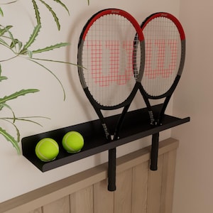 May include: A black metal shelf holding two tennis rackets and two tennis balls. The rackets have red and black frames with red stringing and the Wilson logo. The tennis balls are bright yellow.