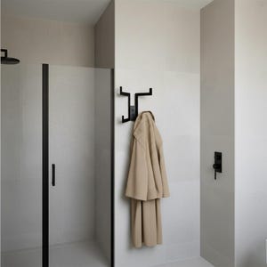 Puede incluir: Un cuarto de baño moderno con una mampara de ducha negra y un perchero negro montado en la pared. Una bata beige cuelga del perchero. Las paredes están alicatadas en un color neutro claro, creando una estética minimalista.