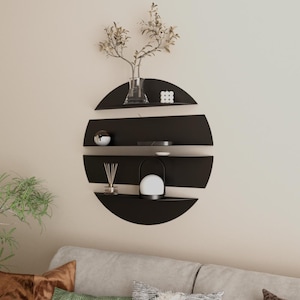 May include: Black, semi-circular wall shelves arranged in a circular pattern, holding decorative items. A vase with dried branches, a small lamp, and other decor pieces are displayed. The shelves are mounted on a neutral-colored wall, with a sofa visible below.