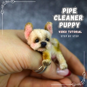 May include: A small pipe cleaner puppy with a beige body, pink ears, and black eyes. The puppy is held in a hand. Text on the image reads "PIPE CLEANER PUPPY VIDEO TUTORIAL STEP BY STEP."