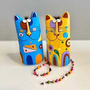 Puede incluir: Dos figuritas de gato de cerámica pintadas a mano con patrones geométricos. El gato azul tiene detalles naranja, rosa y blanco. El gato amarillo tiene detalles marrones, verdes y morados. Un collar de cuentas de colores está drapeado alrededor de los gatos.