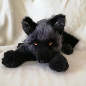 Könnte beinhalten: Ein schwarzer Plüschfuchs mit orangefarbenen Augen und einer weißspitzigen Schwanzspitze. Das handgefertigte Stofftier liegt auf einer weißen Oberfläche, mit ausgestreckten Pfoten. Der Fuchs hat detailliertes Fell und ein realistisches Aussehen.