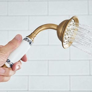 Może przedstawiać: Głowica prysznicowa w stylu vintage z mosiężnym wykończeniem. Uchwyt jest biały z niebieskimi kwiatowymi detalami. Woda wypływa z głowicy prysznicowej, pokazując jej funkcjonalność. Projekt przywodzi na myśl klasyczną, elegancką estetykę.