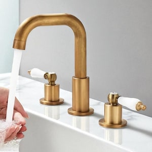 May include: A gold-coloured bathroom tap with a curved spout and two separate handles. The handles have white porcelain accents. Water flows from the tap into a white sink. The tap is a three-piece set.