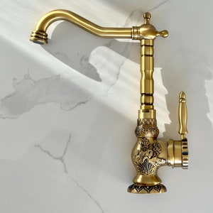 May include: A gold-toned kitchen tap with an ornate design and a single lever handle. The tap has a high spout and a decorative base.