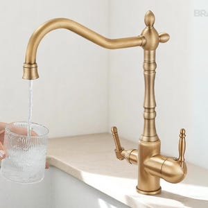 May include: A gold-toned kitchen faucet with a curved spout, filling a clear glass with water. The faucet has a decorative design and is set against a neutral background. The brand name "BRASIFY" is visible in the upper right corner.