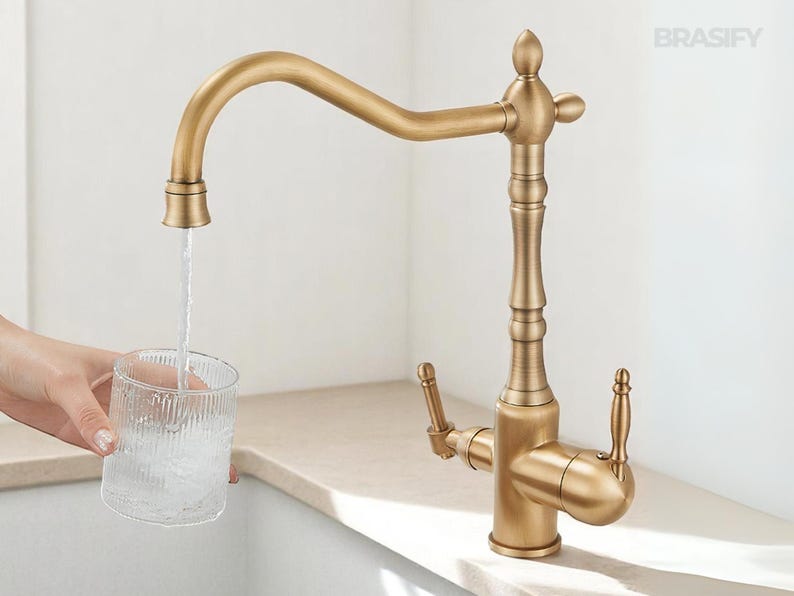 May include: A gold-toned kitchen faucet with a curved spout, filling a clear glass with water. The faucet has a decorative design and is set against a neutral background. The brand name "BRASIFY" is visible in the upper right corner.