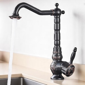 May include: A black kitchen faucet with a curved spout and ornate detailing. Water flows from the faucet into a stainless steel sink. The faucet has a vintage design with a lever handle.