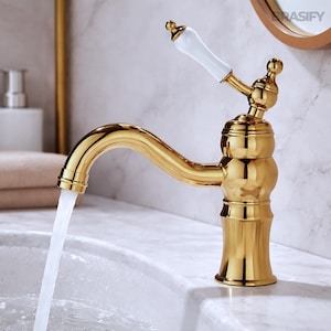 May include: A gold-coloured bathroom tap with a white handle. Water flows from the tap into a white sink. The tap has a classic design with a curved spout and decorative detailing. The tap is made of metal.