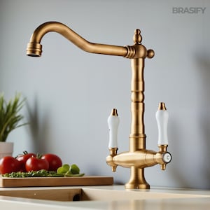 May include: A brass-toned kitchen faucet with a curved spout and white porcelain handles. The faucet has a classic design with a vintage aesthetic. The brand name "BRASIFY" is visible in the upper right corner.