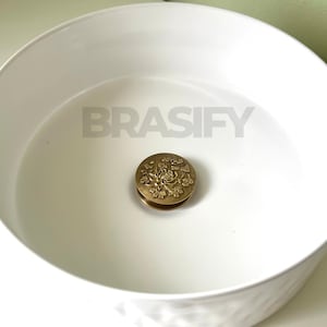 May include: A white ceramic sink with a brass drain stopper featuring a floral design.