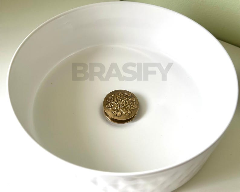 May include: A white ceramic sink with a brass drain stopper featuring a floral design.