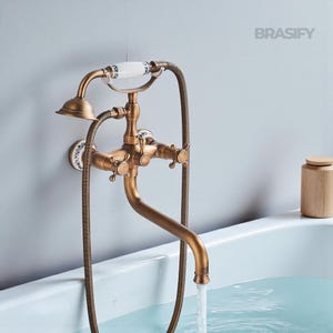 May include: An antique brass-finished wall-mounted bath tap with a handheld showerhead. The tap has white ceramic accents with blue floral patterns and cross-shaped handles. Water flows into a white bathtub.