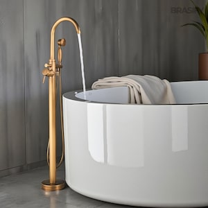Könnte beinhalten: Eine goldfarbene freistehende Badewannenarmatur mit Handbrause. Die Armatur befindet sich neben einer weißen, oval geformten Badewanne. Wasser fließt aus der Armatur in die Wanne. Ein gefaltetes Handtuch liegt am Wannenrand. Der Hintergrund ist eine graue Wand.