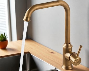 Puede incluir: Un grifo de cocina de color bronce con un caño de cuello de cisne. El agua fluye del grifo a un fregadero de acero inoxidable. El grifo tiene un solo mando de palanca y está montado en una encimera de madera. La marca "BRASIFY" es visible.