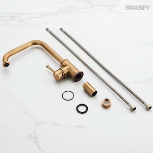 May include: An antique brass kitchen faucet with a curved spout, handle, and various installation components. The faucet is accompanied by two flexible supply lines, a cylindrical piece, and several rings and fittings. The brand name "BRASIFY" is visible in the top right corner.