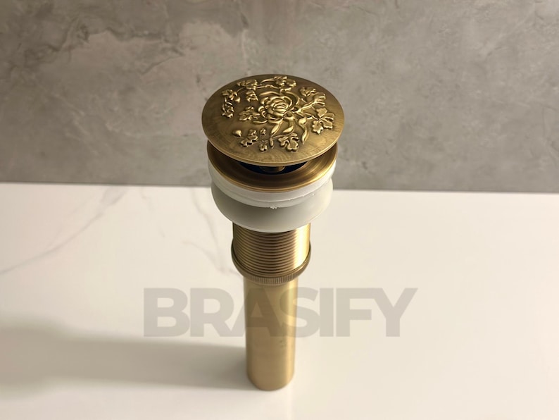 May include: A gold-toned drain stopper with a decorative floral design. The stopper has a white plastic ring around the top and a threaded metal stem.