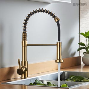 May include: A gold-toned kitchen faucet with a black spring-like hose and a pull-down sprayer. The faucet is mounted above a stainless steel sink, with water flowing into the basin. The faucet has a sleek, modern design.