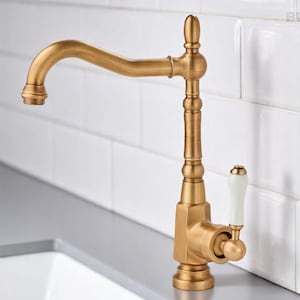 Può includere: Un rubinetto da cucina color oro con un beccuccio curvo e una maniglia bianca. Il rubinetto ha una finitura in ottone antico ed è posto contro un muro piastrellato bianco. Il rubinetto è un design classico.