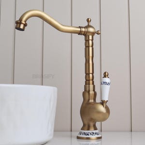 Può includere: Rubinetto da bagno in ottone antico con una leva in ceramica bianca con un motivo floreale blu. Il rubinetto ha un becco alto e sottile e una base decorativa.